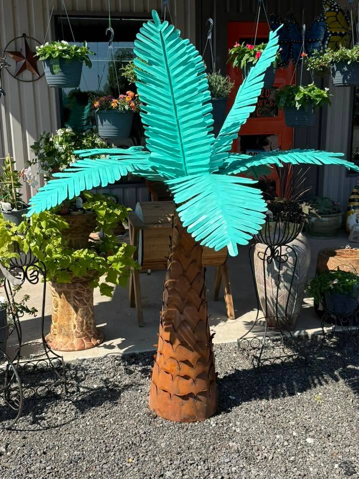 Teal metal palm tree yard art with a rust-colored trunk, surrounded by hanging baskets and potted plants