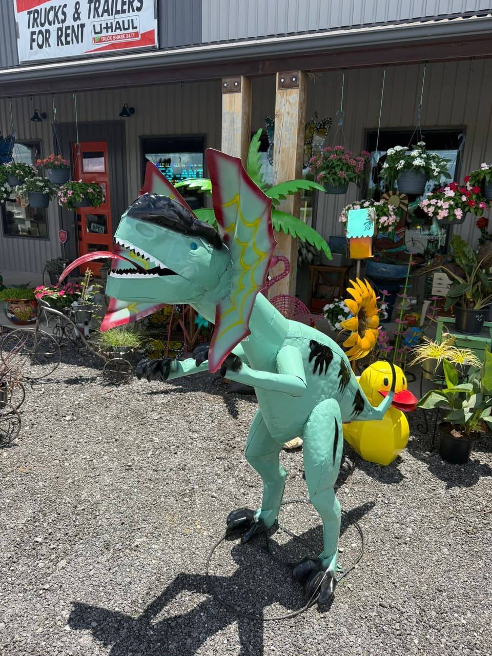Large teal metal dinosaur yard sculpture beside a metal sunflower and a yellow duck on the porch