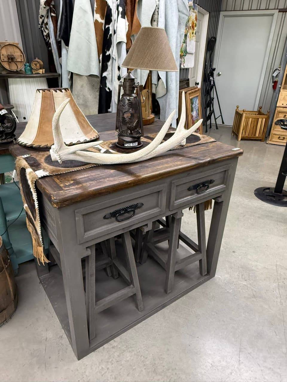 Rustic gray console table with drawers and tuck-away stools, a cowhide runner, a lantern lamp and an antler