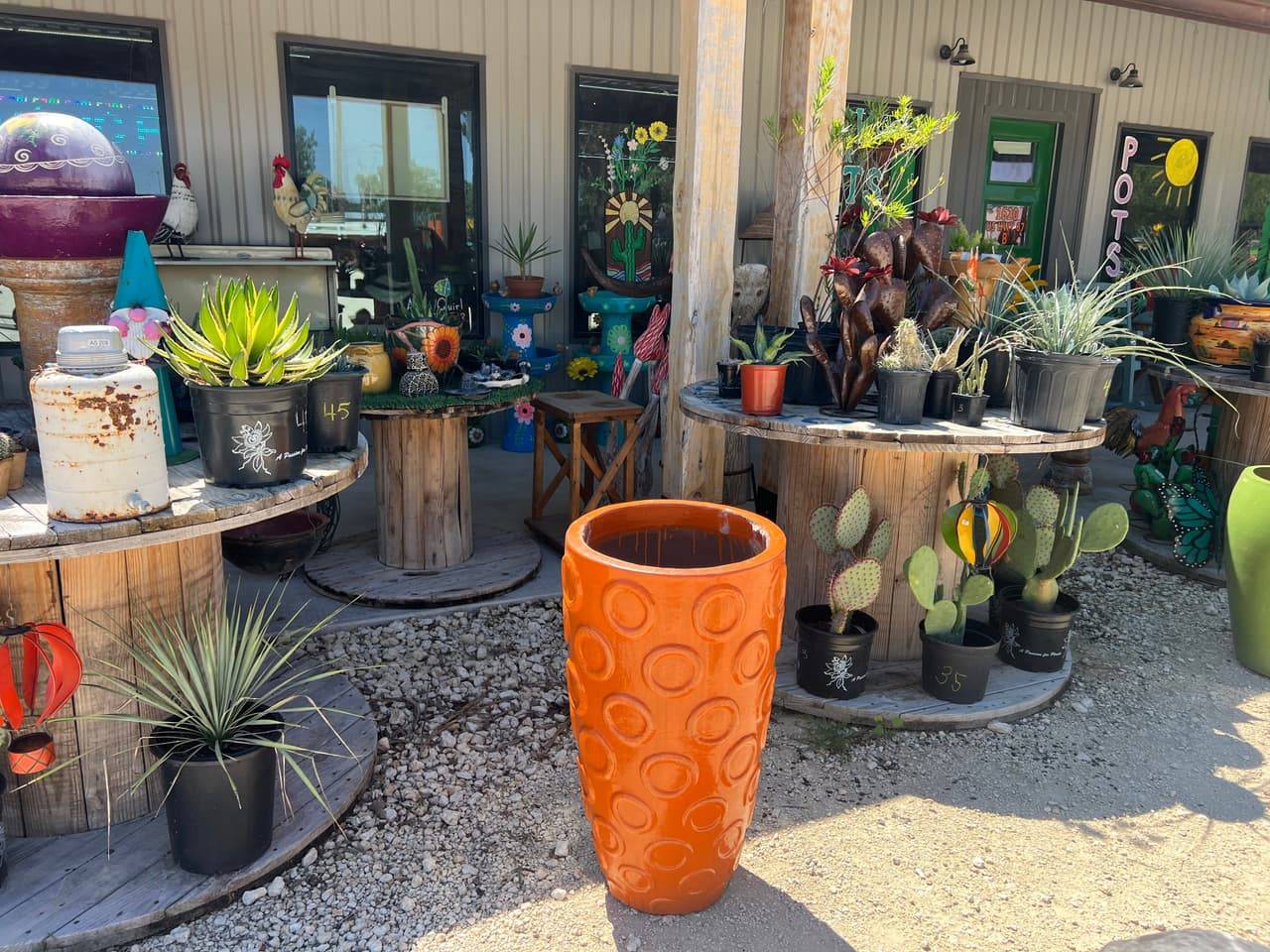 Storefront of Pick Ur Pots in Stephenville, TX with a tall orange glazed pot, cactus metal art and potted plants on cable spool tables